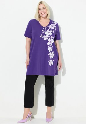 Lilla tunika med V-udskæring og korte ærmer, prydet med hvide blomstermønstre på den ene side; parret med sorte cropped bukser og lavendel højhælede sko.