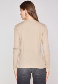 Camicia beige a maniche lunghe con scollatura rotonda e silhouette aderente, abbinata a jeans grigio scuro. Tessuto morbido e dettagli minimali.