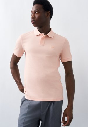 Mand står med hånden på hoften, iført en lyserød kortærmet polotrøje og grå bukser, kigger mod venstre mod en hvid baggrund.