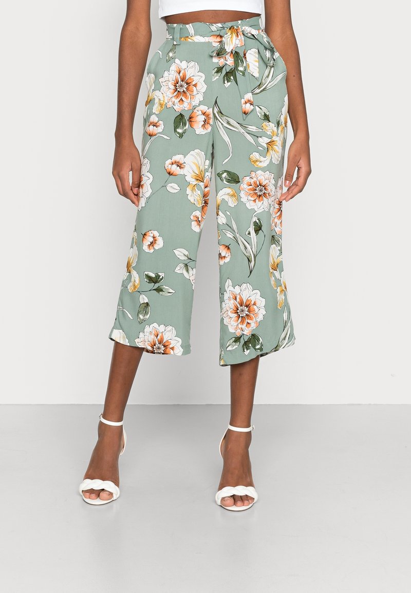 Pantalon large court vert avec imprimé floral orange et blanc, ceinture nouée à la taille haute, porté avec des sandales blanches à bride cheville.