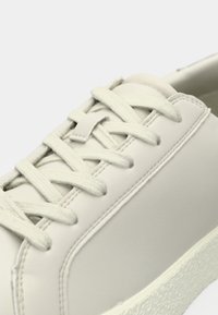 Gros plan d'une basket blanche avec une semelle texturée et des lacets en tissu passés à travers six œillets sur une tige synthétique lisse.