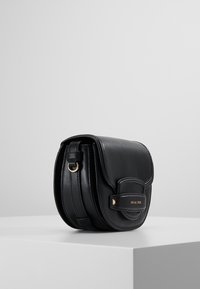 Sac bandoulière en cuir noir avec une forme arrondie, quincaillerie dorée, rabat avant et détail de logo embossé. Texture lisse, design minimaliste.