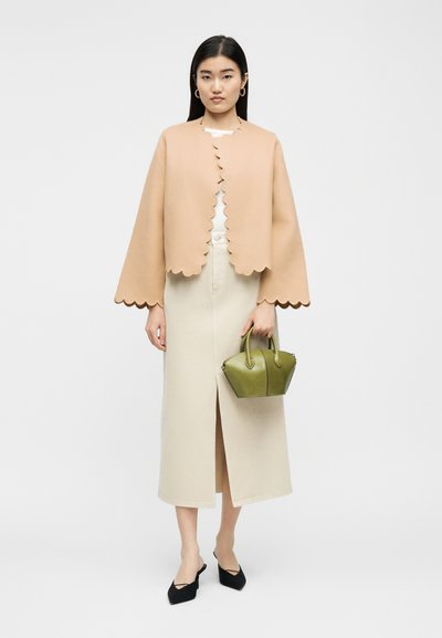 Γυναίκα που φοράει μπεζ σακάκι με κυματιστό τελείωμα, κρεμ φούστα με σκίσιμο μπροστά, μαύρα μυτερά παπούτσια, κρατώντας μικρή πράσινη τσάντα, μπροστά σε απλό φόντο.