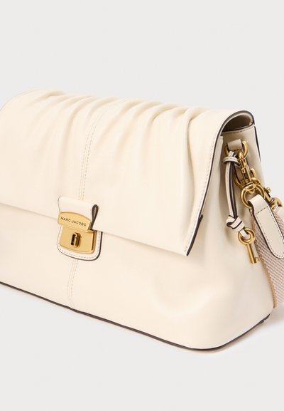 Kremfarvet lædertaske med rynket klap, guldfarvet drejelås, aftagelig rem og nøglering på hvid baggrund.