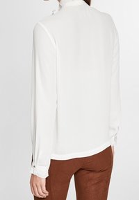 Weiße Bluse mit langen Ärmeln und hohem Kragen, mit geknöpften Manschetten und glatter Textur, kombiniert mit braunen Hosen.