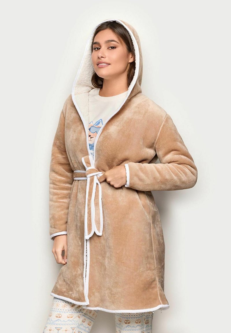 Toga beige con capucha y borde blanco, con una textura suave y un cinturón atado. Llevado sobre pantalones claros con un estampado y una camiseta gráfica.