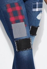 Jean en denim présentant un mélange de patches en tissu à carreaux et unis dans des tons gris, noir et rouge, avec des bords effilochés et des détails usés.