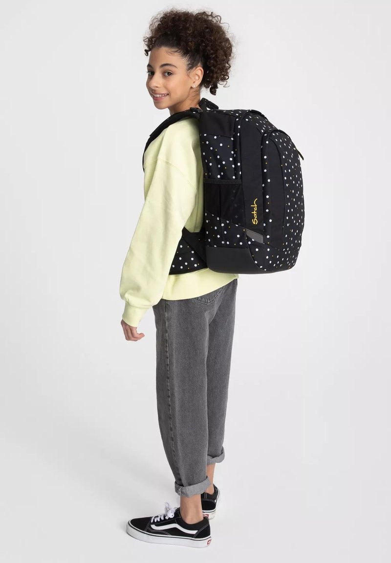 Jeune personne souriante, portant un sweat-shirt jaune clair et un jean gris, portant un grand sac à dos noir avec des points colorés.