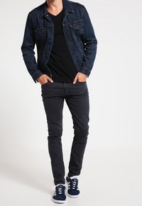 Czarna dżinsowa kurtka na czarną koszulę z dekoltem w serek, w połączeniu z wąskimi czarnymi spodniami i granatowymi sneakersami. Kurtka ma detale z guzikami i kieszenie.