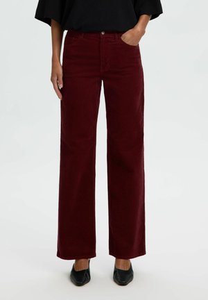 Personne portant un pantalon large rouge foncé avec des chaussures noires, une main dans la poche, debout devant un fond clair uni.