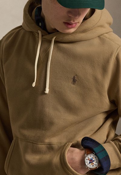 Osoba nosiaca béžovú mikinu s kapucňou a šnúrkami, zelenú čiapku a náramkové hodinky s farebnou postavičkou na ciferníku.