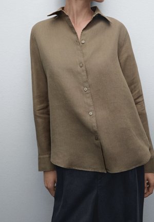 Woman wearing a loose-fitting, buttoned-up olive green shirt with long sleeves and dark pants, standing against a plain light background.