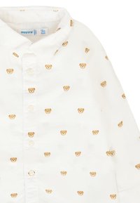 Camicia bianca con bottoni, decorata con piccoli ricami di faccine di orso in tonalità beige e marrone. Presenta un colletto classico e chiusura con bottoni.