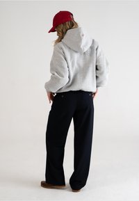 Felpa grigia con cappuccio, dalla vestibilità ampia, con ricami colorati sulla schiena. Abbinata a pantaloni navy a gamba larga e calzature marroni, su uno sfondo neutro.