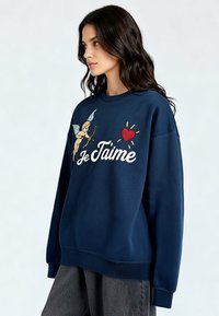 Femme aux longs cheveux bruns portant un sweat-shirt bleu marine avec un cupidon, un cœur rouge, et le texte blanc "Je T'aime", associé à un pantalon gris ample.