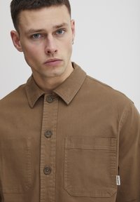 Chemise boutonnière marron en coton, dotée d'un col, de deux poches avant et d'une étiquette logo discrète près de l'épaule. Texture lisse.