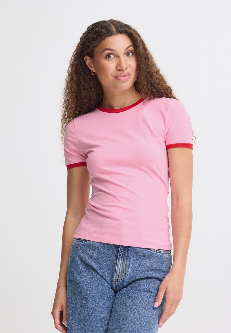 Femme aux cheveux bouclés portant un t-shirt rose avec des bordures rouges et un jean bleu, debout devant un fond blanc uni.