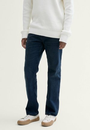 Personne portant un jean bleu foncé, un pull en maille blanc et des baskets blanches avec des semelles beige clair, debout devant un fond uni.