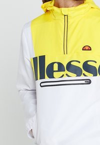 Bright yellow and white windbreaker with a front zip pocket, black "ellesse" logo, and hood. Features a sleek design and smooth texture.