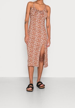 Vrouw die een bruine midi-jurk draagt met bloemenprint, spaghetti-bandjes en een hoge split tot boven het dijbeen, gecombineerd met zwarte sandalen.