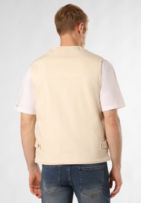 Beige ärmellose Weste aus Baumwollmischung, mit geradem Schnitt, Rückenpasse, Druckknopfverschlüssen und weicher Textur, kombiniert mit einem weißen T-Shirt und blauen Jeans.
