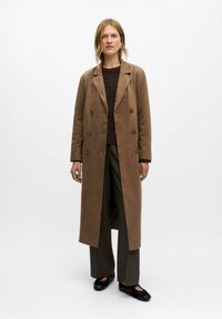 Femme portant un long manteau marron à double boutonnage sur un pull noir et un pantalon large, se tenant devant un fond blanc uni.