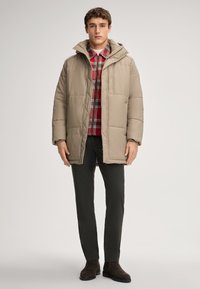 Beżowa kurtka puchowa z kapturem i pikowanym wzorem, noszona na czerwono-czarną koszulę w kratę, w połączeniu z ciemnymi spodniami i brązowymi botkami.