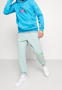 Person klädd i en ljusblå huvtröja med grafik och ljusgröna joggingsbyxor, som intar en dynamisk pose mot en enkel vit bakgrund.