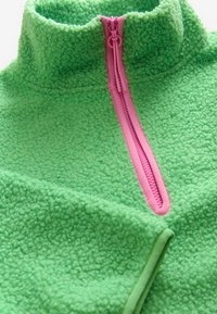 Pullover in pile verde con zip e colletto rosa, caratterizzato da un tessuto a trama e una rifinitura rosa a contrasto lungo la zip.