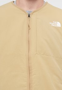 Beige zip-up jakke med rund hals, laget av myk, lett stoff. Har frontlås og logo på venstre bryst.