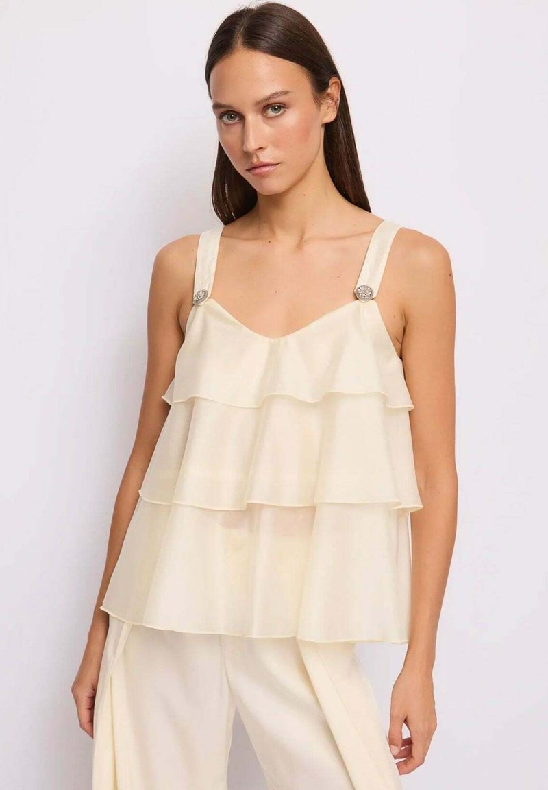 Donna che indossa un top crema senza maniche a balze con bottoni decorativi sulle spalline e pantaloni crema abbinati, in piedi contro uno sfondo semplice.