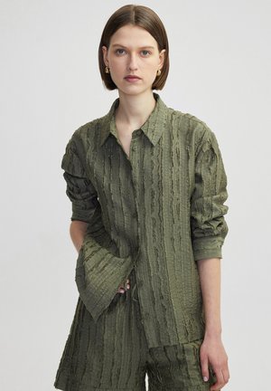 Chemise vert olive en texture, avec un devant à boutons, un col et des manches retroussées. Présente des rayures verticales et une coupe décontractée.