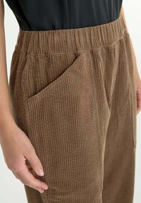 Primer plano de una persona que lleva pantalones de pana marrones con cintura elástica y bolsillos laterales, con la mano descansando cerca del bolsillo.