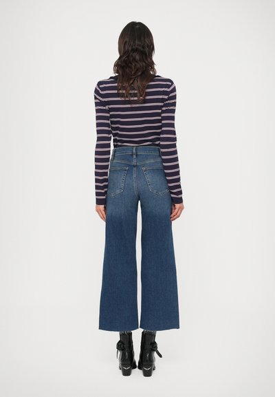 Traper hlače širokih nog z visokim pasom, temno modre barve in z dvema zadnjima žepoma. Kombinirane s prilagojeno črtasto majico z dolgimi rokavi in črnimi gleženjskimi škornji.