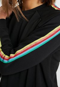 Pull noir à manches longues avec des rayures horizontales jaunes, rouges, bleu clair et vert courant le long des manches, porté par une personne.
