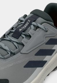 Gray athletic shoe with mesh upper, textured details, and dark blue accents. Features laces and a protective side panel.