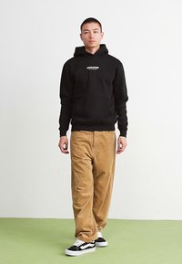 Felpa nera con cappuccio e tasca frontale, pantaloni in corduroy color beige e sneakers bianco e nere. Il modello è in piedi contro uno sfondo neutro.
