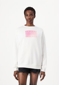 Witte sweatshirt met een ronde hals en lange mouwen, voorzien van een roze gradient logo "TOMMY JEANS" op de voorkant, gecombineerd met zwarte distressed shorts.