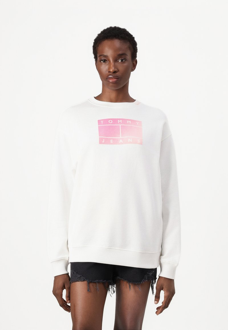 Valge sviiter ümmarguse kaeluse ja pikkade varrukatega, millel on ees roosa gradient logoga "TOMMY JEANS", koos mustade kulunud lühikeste pükstega.