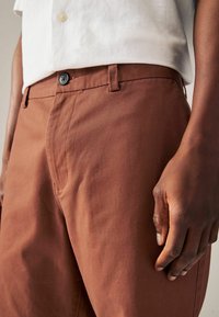 Pantalon en coton marron avec une texture lisse, doté d'une fermeture à bouton et de poches latérales. Gros plan avec une chemise blanche partiellement visible.