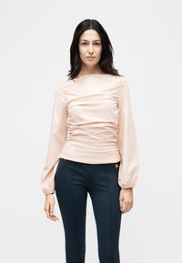 Γυναίκα με ίσια μαύρα μαλλιά, ντυμένη με ανοιχτόροζ μπλούζα με ρυτιδώσεις, μακριά φουσκωτά μανίκια και εφαρμοστά σκούρα μπλε παντελόνια.