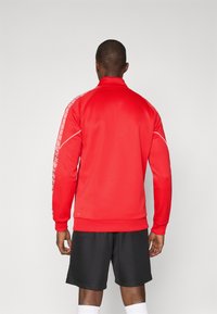 Red zip-up jacket with a high collar, featuring patterned accent stripes along the sleeves and a fitted design. Worn over black shorts.