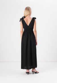 Vestido maxi preto com costas em V, cintura franzida e alças com laço. O tecido parece leve com uma textura suave.