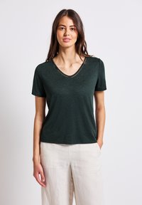 Camiseta de manga corta color verde oscuro con escote en V y pequeños detalles decorativos. Textura suave, corte relajado, combinada con pantalones de lino de color claro.