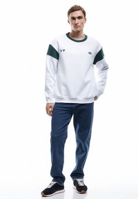 Sudadera blanca con acentos verdes en los hombros, cuello redondo y detalle del logo. Combinada con vaqueros azules y zapatillas negras con cordones blancos.