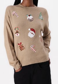 Beżowy sweter dzianinowy z różnymi kolorowymi, cekinowymi motywami świątecznymi: Święty Mikołaj, cukierki, renifery, ozdoby i prezent.