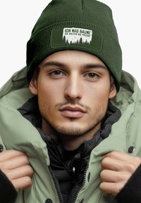 Green knit beanie with a white patch featuring text, worn with a thick, light green jacket layered over a black coat, showcasing zip details.