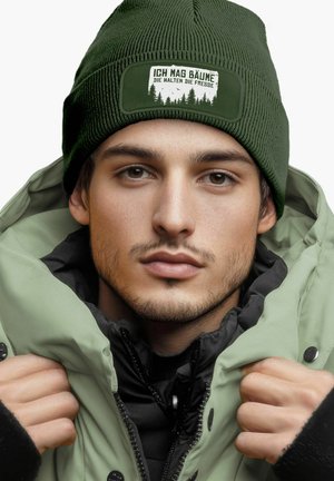 Green knit beanie with a white patch featuring text, worn with a thick, light green jacket layered over a black coat, showcasing zip details.