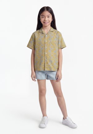 Dievča s dlhými tmavými vlasmi, oblečené v žltej krátkorukávej košeli s vzorom, svetlých riflových kraťasoch a bielych teniskách, stojace a usmievajúce sa.