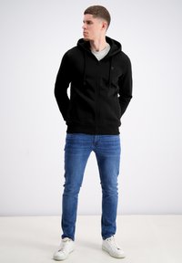 Felpa nera con zip e cappuccio, con lacci, polsini e orlo a coste. Abbinata a jeans blu e sneakers bianche. Sfondo semplice.